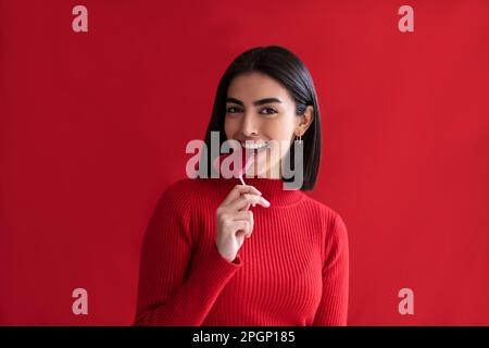 Glückliche junge Frau, die herzförmigen Lutscher vor rotem Hintergrund isst Stockfoto