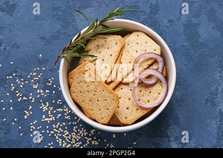 Schüssel mit leckeren Crackern mit Zwiebelringen, Rosmarin und Sesamsamen auf blauem Hintergrund Stockfoto