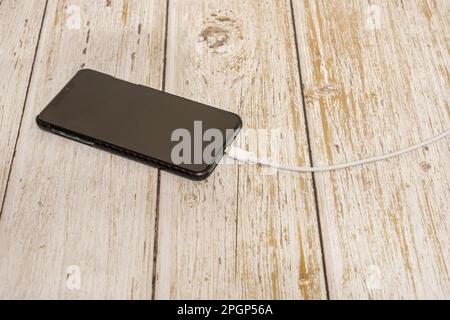 Ein schwarzes Smartphone mit einem weißen Ladekabel, das auf einem Holztisch in das Telefon eingesteckt ist Stockfoto