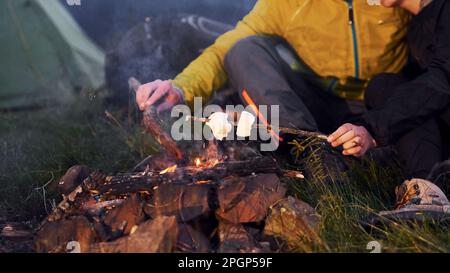 Nahaufnahme der Hände von Menschen, die Stöcke mit Marshmallow im Freien nahe am Lagerfeuer halten Stockfoto