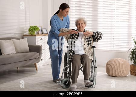 Seniorin im Rollstuhl, die körperliche Bewegung macht, und junge Pflegekraft, die ihr drinnen hilft. Häuslicher Gesundheitsdienst Stockfoto