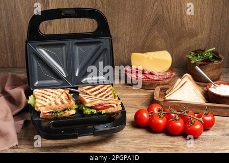 Moderne Grillmaschine mit Sandwiches und verschiedenen Produkten auf Holztisch Stockfoto