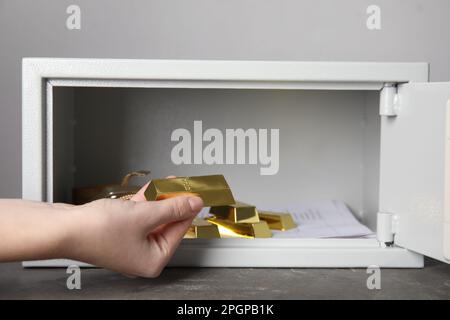 Eine Frau, die Goldbarren aus einem Stahlsafe auf einem grauen Tisch vor hellem Hintergrund nimmt, Nahaufnahme Stockfoto