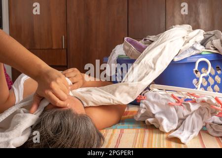 Eine Frau, die auf dem Rücken auf einem Bett liegt, mit einem Haufen Klamotten, den sie neben sich falten kann. Das Faulheitskonzept Stockfoto