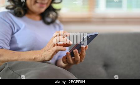 Zugeschnittenes Bild einer glücklichen 60-jährigen, pensionierten Asiatin, die ihr Telefon auf einem Sofa im Wohnzimmer benutzt. Technologie- und Mitarbeiterkonzept Stockfoto