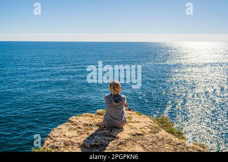 Der Marinha Beach an der Algarve, Portugal, ist das Rückgrat einer jungen Frau, die auf den Klippen sitzt und in die Schönheit des Atlantischen Ozeans eintaucht Stockfoto