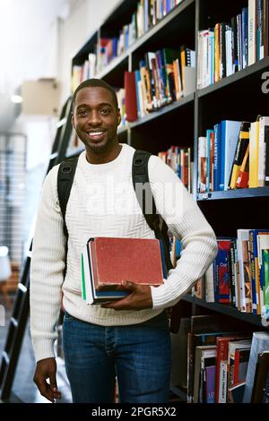 Zeit, die Bücher zu lesen. Porträt eines glücklichen jungen Mannes, der Bücher in einer Bibliothek am College trug. Stockfoto