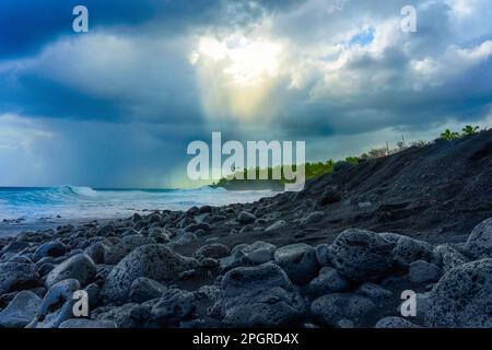 Dramatische Schönheit der hawaiianischen Küste vor einem drohenden Sturm. Die Sonnenstrahlen durchbrechen die Wolken über dem hellblauen Meer und dem schwarzen Sandstrand Stockfoto