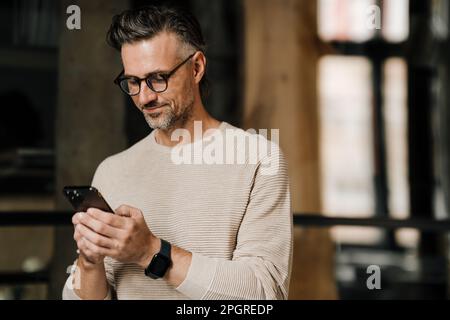 Weißer Mann mittleren Alters mit Brille, der lächelt, während er sein Smartphone im Haus benutzt Stockfoto