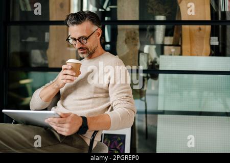 Reifer grauer Mann, der Kaffee trinkt, während er im Büro mit einem Tablet-Computer arbeitet Stockfoto