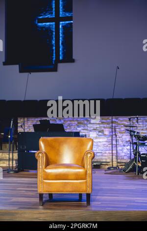 Ein brauner Ledersessel auf der Bühne mit Musikinstrumenten und einem leuchtenden Kreuz im Hintergrund. Stockfoto