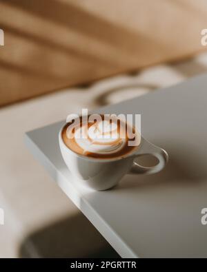 Eine weiße Kaffeetasse aus Keramik gefüllt mit einem dampfenden Latte auf dem Tisch in einer hellen und luftigen Umgebung Stockfoto
