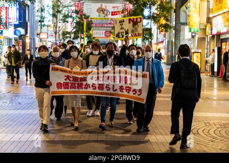 Japanische Demonstranten marschieren am Abend durch eine Einkaufspassage in Kumamoto, mit einem Banner an der Vorderseite und mehreren Demonstranten mit Plakaten. Die Demonstration ist gegen eine Erhöhung der Ausgaben für die Zivilschutztruppe. Stockfoto