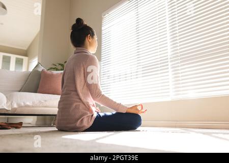 Blick aus dem niedrigen Winkel auf eine weiße junge Frau, die vor Fensterläden meditiert, während sie auf dem Boden sitzt. Kopierraum, unverändert, Yoga, Zen, Lifestyle, Exer Stockfoto