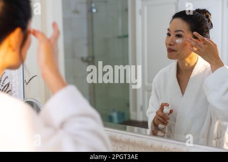 Lächelnde asiatische Frau im Bademantel, die vor dem Spiegel steht und Gesichtscreme im Badezimmer aufträgt. Lifestyle, Selbsthilfe, Gesundheit und Schönheit. Stockfoto