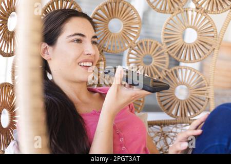 Eine weiße, glückliche junge Frau, die über den Lautsprecher über ein Smartphone spricht, während sie zu Hause in der Schaukel sitzt. Unveränderte Technologie, Lifestyle, Kommunikation und rel Stockfoto