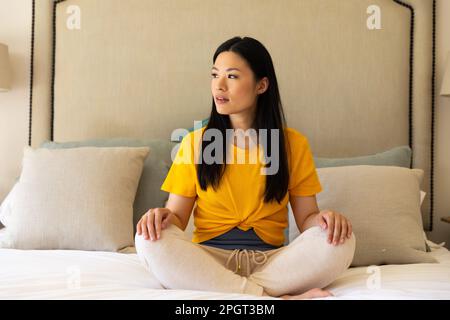 Rücksichtsvolle asiatische Frau, die quer auf dem Bett sitzt, meditiert, wegschaut. Gesunder Lebensstil, Freizeit und Entspannung. Stockfoto