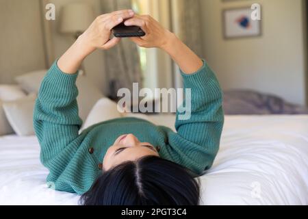 Lächelnde asiatische Frau, die zu Hause im Bett lag und ein Smartphone benutzte. Lifestyle, Freizeit, Kommunikation und Entspannung. Stockfoto