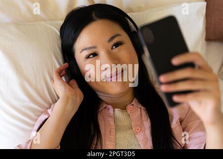 Lächelnde asiatische Frau, die auf dem Bett liegt und Kopfhörer mit Smartphone trägt. Lifestyle, Freizeit, Kommunikation und Entspannung. Stockfoto
