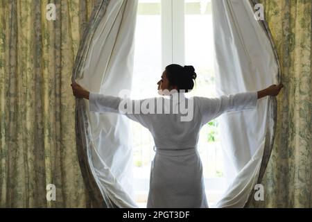 Glückliche Asiatin im Bademantel, die morgens die Vorhänge im Schlafzimmer öffnete. Häusliches Leben, Freizeit und Entspannung. Stockfoto