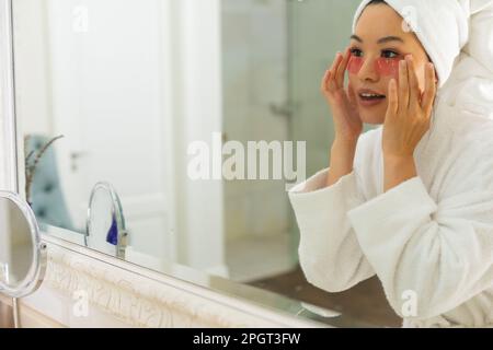 Eine glückliche Asiatin im Bademantel, die in den Badezimmerspiegel guckt und Augenklappen aufträgt. Lifestyle, Selbsthilfe, Gesundheit und Schönheit. Stockfoto