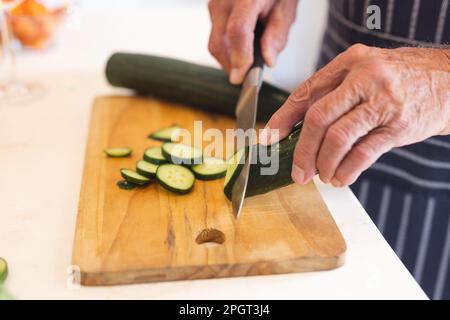 Die Hände eines weißen Seniors, der eine blaue Schürze trägt und Gurken schneidet, während eines Kochkurses. Ruhestand, Lernen, Kochen und gesunder Lebensstil. Stockfoto