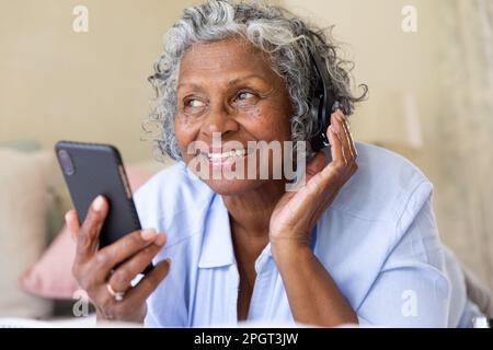 Lächelnde afroamerikanische Seniorin, die Musik hört und ihr Smartphone benutzt, während sie im Bett liegt. Unverändertes, Lifestyle, Entspannung, Technologie, Kopfhörer, Stockfoto