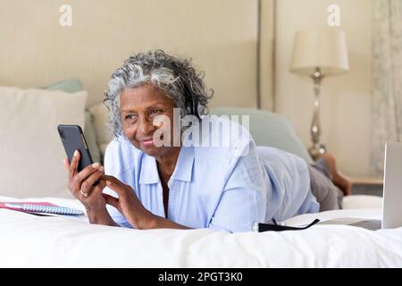 afroamerikanische Seniorin, die Musik über Kopfhörer hört und Handy benutzt, während sie im Bett liegt. Kopierraum, unverändert, Lifestyle, Entspannung, Technik Stockfoto
