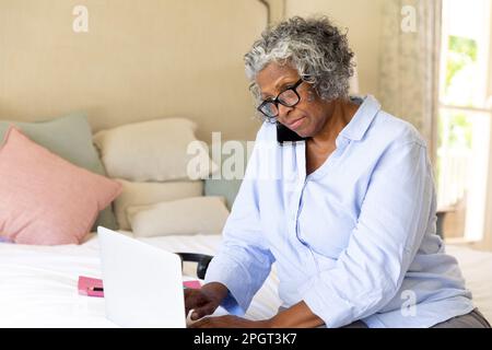 afroamerikanische Geschäftsfrau, die sich auf ihre Bedürfnisse konzentriert und über ein Smartphone spricht und ein Notebook im Bett benutzt. Kopierraum, unverändert, professionell, Arbeit von zu Hause aus, EY Stockfoto