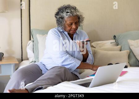 Eine afroamerikanische Geschäftsfrau, die sich über Handylautsprecher unterhält und ein Notebook auf dem Bett benutzt. Kopierbereich, unveränderter, professioneller, Arbeit von zu Hause aus, voi Stockfoto