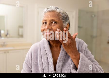 Porträt einer afroamerikanischen Seniorin im Bademantel, die zu Hause Gesichtscreme aufträgt. Unverfälscht, Körperpflege, Altern, Verwöhnung, Porträt, Beauty PR Stockfoto