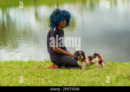 Goiania, Goias, Brasilien – 20. März 2023: Eine junge schwarze Frau mit gefärbtem blauem Haar sitzt mit ihren Hunden auf dem Rasen neben einem See. Stockfoto