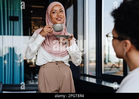 Muslimische junge Geschäftsfrau, die Kaffee trinkt, während sie mit ihrer Assistentin im Büro spricht Stockfoto