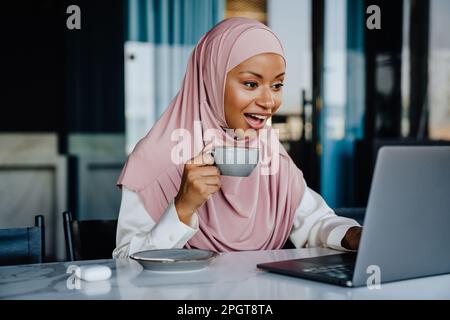 Muslimische junge Geschäftsfrau, die Kaffee trinkt, während sie im Büro an einem Laptop arbeitet Stockfoto