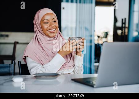 Muslimische junge Geschäftsfrau, die Kaffee trinkt, während sie im Büro an einem Laptop arbeitet Stockfoto