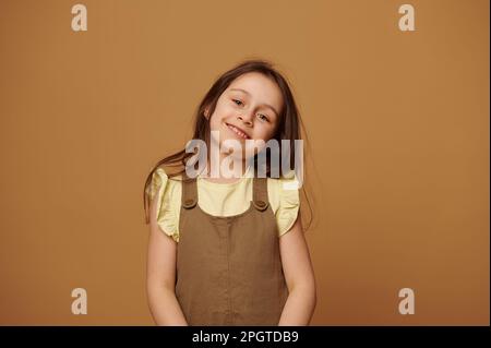 Ein bezauberndes kleines Mädchen, das lächelt und in die Kamera schaut, isoliert auf beigem Hintergrund. Wunderschöne Kinder. Kind im Grundalter Stockfoto