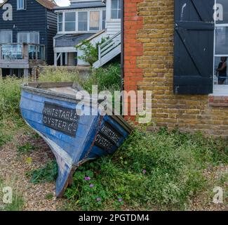 Wetterbestes Holzkanu mit Whitstable Oyster Co auf beiden Seiten, auf Laub neben einem Backsteingebäude am Whitstable Beachfront. Stockfoto