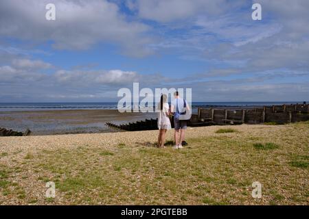 Mann und Frau stehen am Kieselstrand mit Blick auf das Meer neben der hölzernen Groyne. Whitstable West Beach, Nordost-Kent Coast, England, Großbritannien. Stockfoto