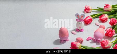 Rosafarbene Tulpen mit Ostereiern und Kaninchen auf hellem Steinbetonhintergrund. Festliches Konzept, Grußkarte, flach liegend, hartes Licht, dunkler Schatten, Banner Stockfoto