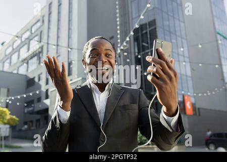 Glücklicher afroamerikanischer Geschäftsmann, der per Videoanruf mit seinem Team spricht. Sie stehen vor einem Bürozentrum, tragen Kopfhörer und halten ein Telefon in der Hand. Stockfoto