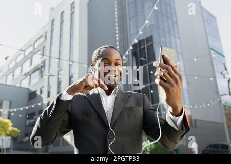 Ein älterer schwarzer Mann im Anzug steht vor einem Business Center und telefoniert per Videoanruf über Kopfhörer. Stockfoto