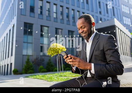 Lächelnder Geschäftsmann aus Afroamerikanern im Anzug, sitzt auf einer Bank außerhalb des Büros, trägt Kopfhörer und spricht am Telefon. Stockfoto