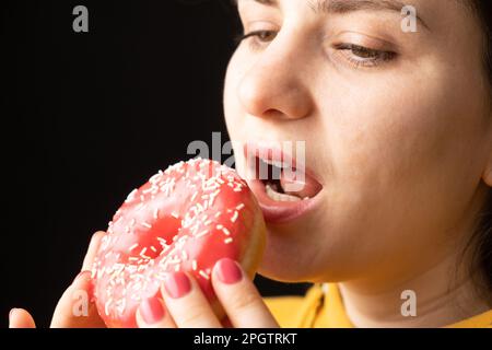 Eine Frau beißt einen großen roten Donut, einen schwarzen Hintergrund, ein Ort für Text. Gluttonie, überfressen und zuckersüchtig. Stockfoto