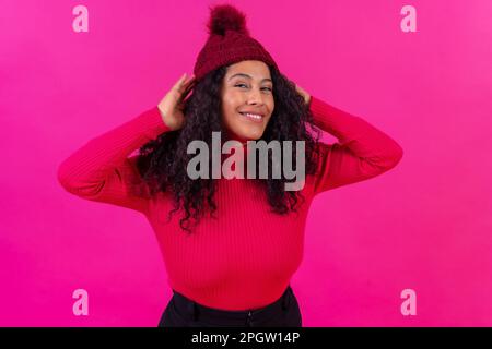 Eine fröhliche hispanische Frau mit lockigem Haar in einem Hut auf einem leeren pinken Hintergrund Stockfoto