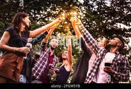 Schicke Jahrtausendleute, die Spaß mit Feuerglitzern haben und Wein toasten - Junge, trendige Freunde jubeln auf einer Picknick-Barbecue-Party bei nächtlicher Stimmung draußen Stockfoto