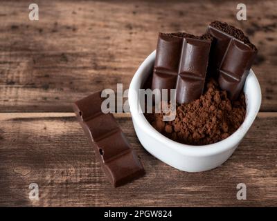 Bittere Schokoladenriegel mit Porenbildung und Kakaopulver in weißer Schüssel auf dem Holztisch, selektiver Fokus. Stücke dunkelporöser Schaumschokolade Stockfoto