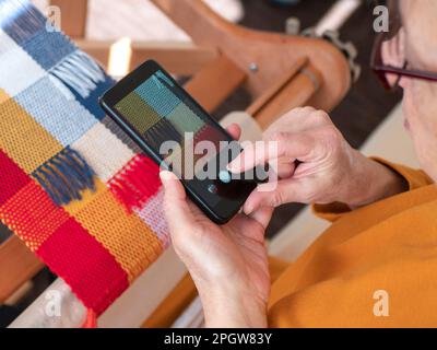 Finger eines Handwerkers, der den Bildschirm eines Mobiltelefons berührt, um ein Bild von ihrem Weberei-Projekt zu machen, Draufsicht. Drücken der Kamerataste, selektiver Fokus Stockfoto