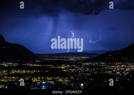 Ein Gewitter erhellt den Nachthimmel über einer Metropolregion mit einer weit entfernten Bergkette im Hintergrund Stockfoto