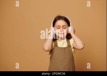 Das fröhliche kleine Mädchen hört den Soundtrack über die Kopfhörer, hört beruhigende Musik mit geschlossenen Augen und beigefarbenem Hintergrund Stockfoto