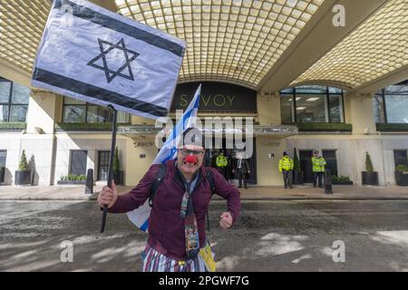 London, Großbritannien. März 2023. Ein Demonstrant in satirischen Kostümen, einschließlich einer roten Clownnnase und einem Helm, posiert energisch mit einer israelischen Flagge vor dem Eingang des Savoy Hotels in London. Sicherheitspersonal in gut sichtbaren Jacken überwachen den Tatort. Penelope Barritt/Alamy Live News Stockfoto